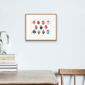 May include: A framed print of a colorful illustration featuring 12 cartoon characters with different expressions and outfits. Each character is a round, orange shape with a face and two small wings. The characters are arranged in a grid pattern.