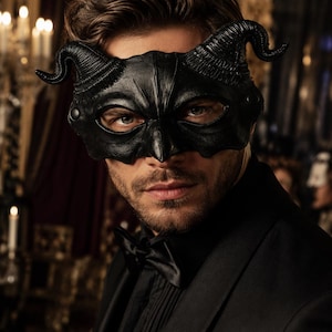 Può includere: Una maschera da ballo in maschera nera con due corna ricurve sui lati e dettagli strutturati. La maschera copre la parte superiore del viso, compresi gli occhi, ed è indossata con una giacca da abito nera e un papillon nero abbinato.