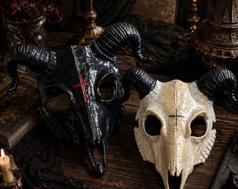 May include: Two animal skull masks with curved horns. The left mask is glossy black with a red cross symbol on the forehead. The right mask is off-white with a textured bone finish and a small black cross symbol between the eye sockets.