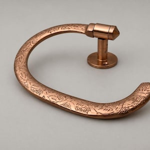 May include: A copper-toned metal towel ring featuring a curved, open-ended design with an embossed grape and vine pattern. The fixture includes a circular wall-mounting plate and a cylindrical arm that connects to the main ring structure.