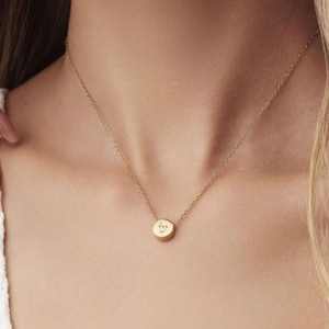 May include: A gold-toned necklace featuring a delicate chain and a small, circular pendant. The pendant is engraved with a single Hebrew letter, specifically the character Shin, centred on its smooth, polished surface. The chain is thin and metallic.