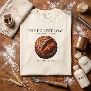 Op de afbeelding: Een crèmekleurig t-shirt met een afbeelding van een rond brood. De tekst luidt THE HONEST LOAF, Flour Water Salt Time en NOTHING ADDED. Het t-shirt heeft een ronde hals en korte mouwen en ligt op een houten ondergrond.
