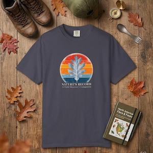 May include: A charcoal grey short-sleeved t-shirt featuring a graphic of a white oak leaf centred within a circular sunset design of red, orange and blue stripes. Below the graphic, the text reads "NATURE'S RECORD A Field Observer's Companion."