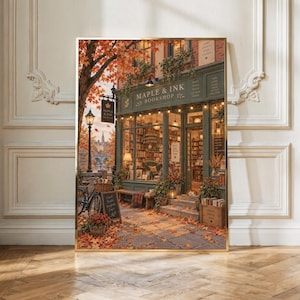 May include: A framed wall art print depicting a cosy bookstore storefront named Maple & Ink Bookshop. The scene features autumn foliage, a bicycle, a chalkboard sign, and warm interior lighting visible through the large glass windows.