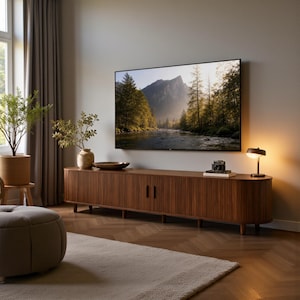 May include: A long, dark wood media console with vertical slats and rounded ends, positioned against a wall below a large flat-screen television displaying a mountain landscape. A small lamp, a camera, and a vase with greenery sit on top.