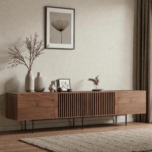 May include: A long, low-profile wooden media console featuring two solid wood end cabinets and two central slatted doors. The unit is supported by thin black metal legs and is topped with several ceramic vases and small framed art pieces.