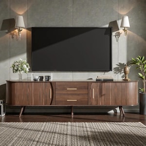 May include: A long, rectangular wooden media console featuring two central drawers with gold-toned horizontal handles, flanked by two curved, slatted cabinet doors. The unit stands on tapered wooden legs and is positioned beneath a large flat-screen television.