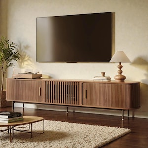 May include: A long, wooden media console with rounded edges, vertical slat detailing, and two cabinet doors with thin metal handles. A flat-screen television is mounted on the wall above the console, which holds a table lamp and books.