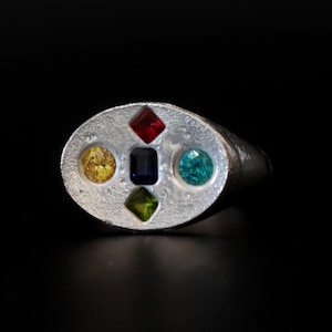 May include: A silver-toned ring featuring an oval-shaped face with a textured surface. The face is set with five gemstones: a red diamond-cut stone at the top, a green diamond-cut stone at the bottom, a yellow round stone, a blue round stone, and a central dark blue rectangular stone.