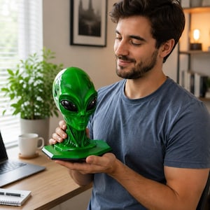 Puede incluir: Una escultura de cabeza alienígena de color verde brillante y lustroso, con ojos ovalados grandes, lisos y negros, y una estructura de cuello definida. La cabeza está montada sobre una base hexagonal plana, con un acabado vibrante y detallado.
