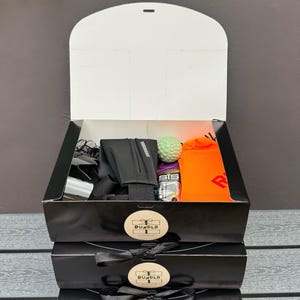 Puede incluir: Una caja de regalo negra con interior blanco que contiene un cinturón de correr negro, una pelota de masaje texturizada de color verde claro, un sobre de gel energético SiS y un par de calcetines de color naranja brillante. Tiene una etiqueta BUNDLD.