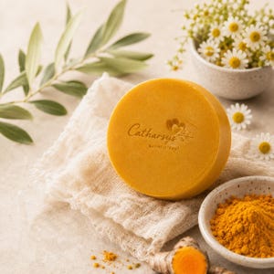 May include: A round, golden-yellow bar of soap featuring the embossed text Catharsys holistic soul. The soap rests on a textured, off-white fabric cloth, accompanied by a small bowl of yellow turmeric powder and a fresh turmeric root piece.