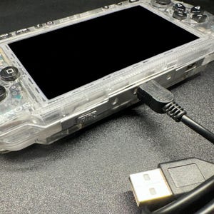 Könnte beinhalten: Eine durchsichtige, transparente Handheld-Spielkonsole mit schwarzem Bildschirm und schwarzen Tasten, die auf der linken Seite ein Sony-Logo aufweist. Ein schwarzes Ladekabel ist an der Seite des Geräts angeschlossen, mit einem USB-Stecker am Ende.