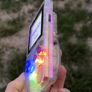 Può includere: Una console di gioco portatile Game Boy Color con scocca trasparente. Il dispositivo presenta uno schermo che mostra il testo GAME BOY, un pad direzionale blu e componenti interni illuminati da luci LED blu, gialle e rosse.