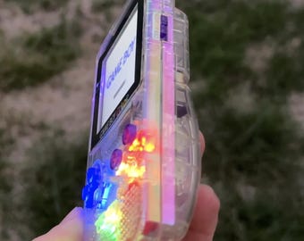 Peut inclure: Une console de jeu portable Game Boy Color avec une coque transparente. L'appareil dispose d'un écran rétroéclairé affichant le texte GAME BOY, d'une croix directionnelle bleue et de composants internes illuminés par des lumières bleu, jaune et rouge.