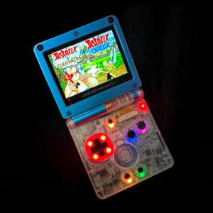 Pode incluir: Uma consola portátil Game Boy Advance SP azul com caixa transparente e botões iluminados. O ecrã apresenta o título Asterix & Obelix, com personagens de desenhos animados coloridas. O dispositivo tem um D-pad e botões de ação.