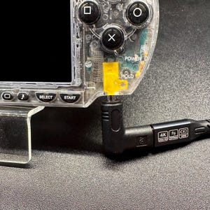 Op de afbeelding: Een draagbare spelcomputer van doorzichtig plastic met zwarte knoppen en een geel intern onderdeel, verbonden met een zwarte L-vormige oplaadkabel. De kabelconnector toont iconen voor 4K Ultra HD, 20Gbps en 240W vermogen.