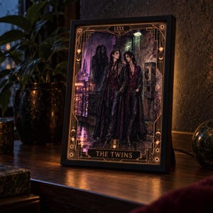 Könnte beinhalten: Ein rechteckiger Leuchtkasten mit schwarzem Rahmen zeigt eine beleuchtete Illustration im Tarot-Stil mit dem Titel THE TWINS. Das Bild zeigt zwei Figuren in dunkler, mehrlagiger Kleidung in einer regnerischen, düsteren Stadtkulisse.