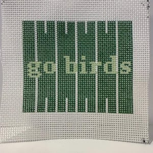 May include: A square piece of white needlepoint canvas featuring a centered design of vertical dark green stripes resembling a football field. The text go birds is stitched in a light green color across the center of the striped pattern.