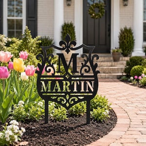 May include: A black metal garden sign featuring a decorative shield shape with a fleur-de-lis top, a large letter M, and the name MARTIN cut out in the center. The sign is supported by two thin metal stakes inserted into the dark soil of a garden bed.