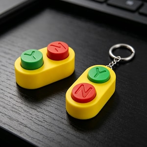 Może przedstawiać: Dwa żółte, owalne urządzenia typu fidget z wypukłymi przyciskami. Jedno z nich to brelok z metalowym kółkiem. Każde urządzenie posiada zielony przycisk z literą Y oraz czerwony przycisk z literą N, ułożone w pary.