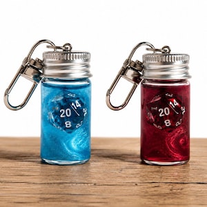 Puede incluir: Dos llaveros de frasco de vidrio, uno lleno de líquido azul brillante y el otro con líquido rojo brillante. Cada frasco contiene un dado de veinte caras con números blancos, incluidos el 20, 14 y 8, visibles a través de las paredes de vidrio.