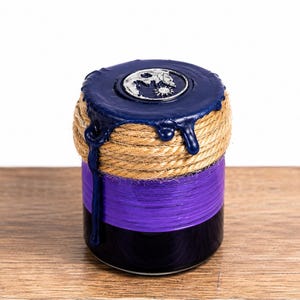 Puede incluir: Un frasco cilíndrico con base negra y una sección central morada envuelta en cuerda. La tapa está sellada con cera azul oscuro que gotea por los lados, con un emblema circular plateado que representa un diseño de sol y luna en la parte superior.