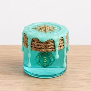 May include: A clear glass jar filled with teal liquid containing a twenty-sided die with gold numbers. The lid is sealed with light teal wax that drips down the sides, wrapped with natural brown twine and topped with a gold compass emblem.
