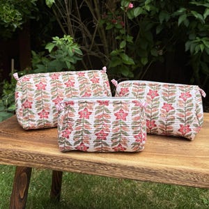 Peut inclure: Trois trousses matelassées rectangulaires présentant un motif floral répétitif avec des fleurs roses et des feuilles vertes sur fond blanc. Chaque trousse possède une fermeture éclair et de petites languettes en tissu sur les côtés.
