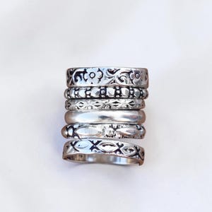 Könnte beinhalten: Ein Stapel aus sechs silberfarbenen Metallringen mit verschiedenen Texturen und Mustern. Die Designs umfassen florale Motive, Perlenverzierungen, geometrische Formen und ein glattes, poliertes Band, die alle vertikal in einem Set angeordnet sind.