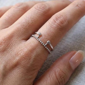 Könnte beinhalten: Ein silberfarbener offener Ring mit doppeltem Banddesign. Das untere Band ist mit kleinen, gleichmäßig verteilten Perlen texturiert, während der obere Teil zwei vertikale Stifte aufweist, die jeweils mit einem kleinen, runden Blumenakzent versehen sind.