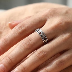 Könnte beinhalten: Ein silberfarbener Metallring mit einem komplizierten, durchbrochenen Filigranmuster aus geschwungenen, rankenartigen Motiven, die sich um das gesamte Band ziehen. Das Design hat ein dunkles, oxidiertes Finish in den Vertiefungen.