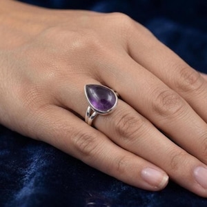 Könnte beinhalten: Ein Silberring mit einem tropfenförmigen, violetten Amethyst-Edelstein, der in ein poliertes Metallband eingefasst ist. Der Ring verfügt über ein geteiltes Schienendesign, das mit der Fassung verbunden ist und eine glatte, abgerundete Steinoberfläche zeigt.