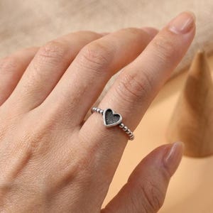 Könnte beinhalten: Ein silberfarbener Ring mit einem kleinen herzförmigen Mittelstück mit einer dunklen, strukturierten Einlage. Das Ringband besteht aus einer Reihe kleiner, miteinander verbundener Metallkugeln, die ein perlenartiges Aussehen um den Finger erzeugen.
