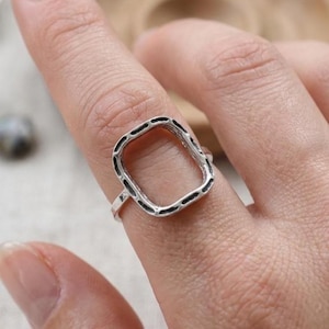Könnte beinhalten: Ein silberfarbener Ring mit einem großen, offenen rechteckigen Rahmen. Die Metalloberfläche des Rings hat eine texturierte, leicht abgedunkelte Oberfläche mit kleinen Vertiefungen entlang des Umfangs der rechteckigen Form, was einen deutlichen visuellen Kontrast erzeugt.