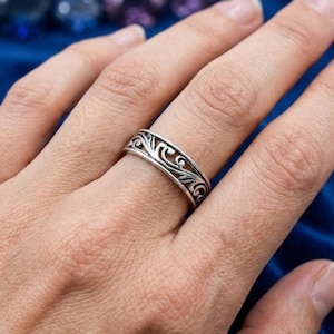 Könnte beinhalten: Ein Ring aus silberfarbenem Metall mit einem komplexen, durchgehenden durchbrochenen Schnörkelmuster, das sich um das gesamte Band zieht. Das Design besteht aus ineinandergreifenden geschwungenen Linien und Kurven mit strukturierter Oberfläche.