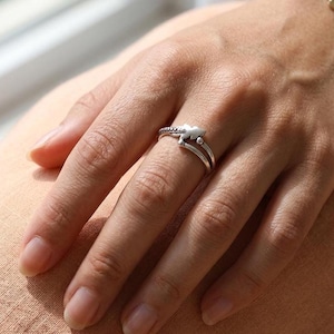 Könnte beinhalten: Ein silberner Ring mit einem geteilten Banddesign, das in einem zentralen Punkt zusammenläuft. Das Metall hat eine polierte Oberfläche mit einem dezenten, texturierten Detail an einer Seite des Bandes, was eine geschichtete Optik erzeugt.