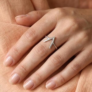 Könnte beinhalten: Ein silberfarbener Ring mit einem V-förmigen Design und einer spitzen Oberseite. Die Ringschiene ist dünn und glatt. Das Metall hat eine matte Oberfläche, und die geometrische Form erzeugt eine markante, kantige Silhouette am Finger.