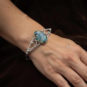 Könnte beinhalten: Ein silbernes Armband mit einem ovalen Stein, der ein mosaikartiges Muster aus blauen, grünen und dunkelbraunen Segmenten aufweist. Das Metallband zeigt auf beiden Seiten der zentralen Steinfassung dekorative, durchbrochene Schnörkel.