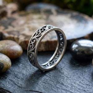Könnte beinhalten: Ein silberfarbener Metallring mit einem komplizierten, durchbrochenen Filigranmuster aus wirbelnden Schnörkeln und Perlendetails. Das Band weist ein dunkles, oxidiertes Finish in den vertieften Bereichen auf, um das Relief hervorzuheben.
