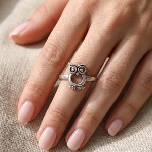 Könnte beinhalten: Ein Ring aus silberfarbenem Metall mit einem stilisierten Eulenmotiv. Das Eulen-Design ist mittig auf der Ringschiene platziert und zeigt kreisförmige Augen, einen kleinen Schnabel sowie einen hohlen, runden Körper. Das Metall hat ein oxidiertes Finish.
