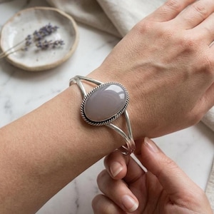 Peut inclure: Un bracelet manchette en argent orné d'une grande pierre ovale grise lisse sertie dans un cadre en métal torsadé. Le bracelet présente un design à double tige avec deux bandes en argent poli qui se rejoignent au niveau du sertissage central.