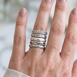 Puede incluir: Un conjunto de cinco anillos de plata apilados en un dedo. Incluye una banda trenzada, una banda con grabado floral, una banda lisa pulida, una banda con textura ondulada y una banda con un patrón geométrico repetitivo en acabado oxidado.