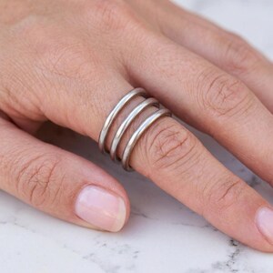 Könnte beinhalten: Ein Set aus drei dünnen, glatten, silberfarbenen Metallringen, die zusammen an einem Finger getragen werden. Die Bänder haben ein abgerundetes Profil und eine polierte Oberfläche, was einen gestapelten Look erzeugt. Jeder Ring ist identisch.