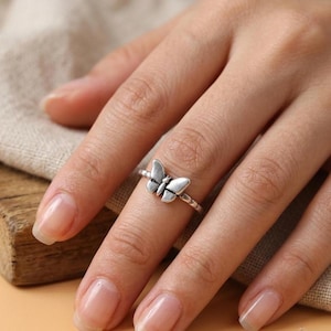 Könnte beinhalten: Ein Ring aus silberfarbenem Metall mit einem schmetterlingsförmigen Mittelstück. Die Ringschiene hat eine texturierte, seilartige Optik, und das Schmetterlingsdesign weist oxidierte Details in den Vertiefungen auf, um die Flügel hervorzuheben.