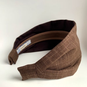 May include: A brown fabric headband with a knotted detail on one side. The headband is made of a textured fabric and has a brown elastic band on the inside.