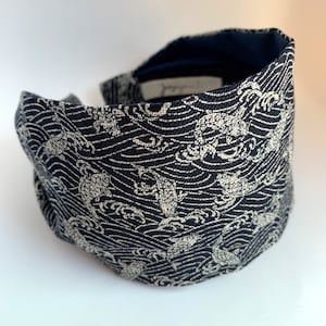 May include: A black headband with a white pattern of waves and turtles. The headband is made of a soft, woven fabric.