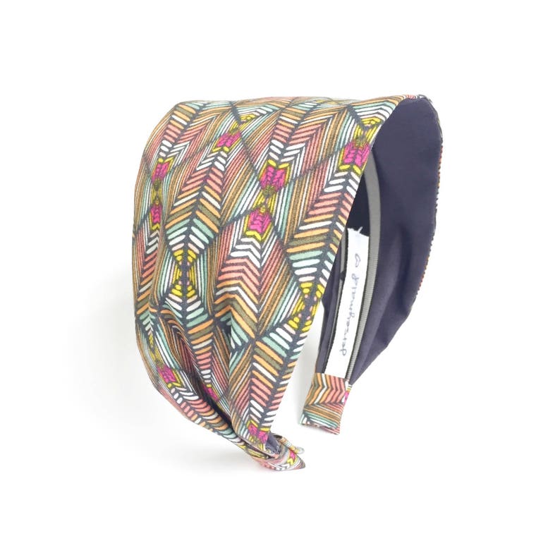 May include: A wide headband with a colorful geometric pattern of feathers in shades of pink, orange, yellow, and teal. The interior is a solid navy blue, and a small tag with text is visible. The headband is designed for comfort and style.