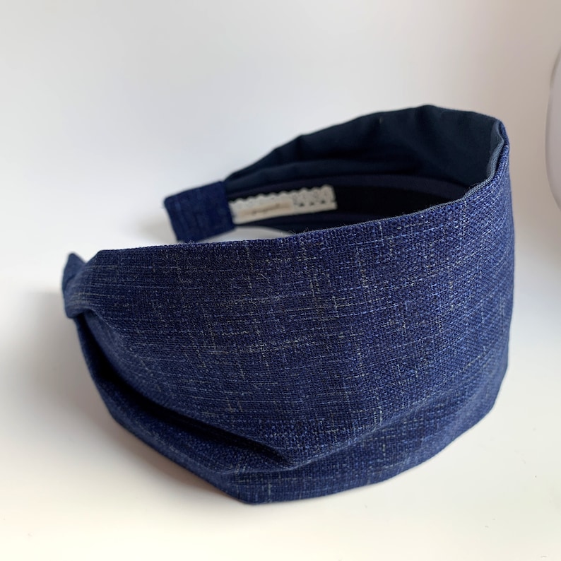 May include: A dark blue fabric headband with a knotted design. The headband is made of a textured fabric.