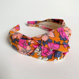 Op de afbeelding: Een geknoopt hoofdband met een bloemenprint in tinten oranje, roze en blauw. De hoofdband is gemaakt van stof en heeft een gedraaid knoopontwerp.